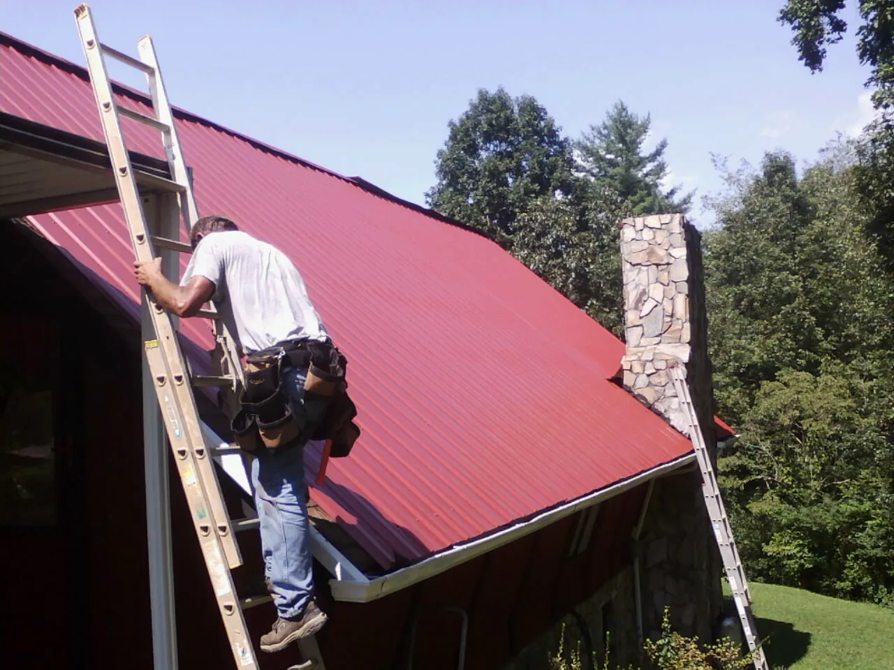Roofer on a ladder beside a red metal roof and stone chimney — Soffit & Fascia Repair by Roofing Brothers in Countyline